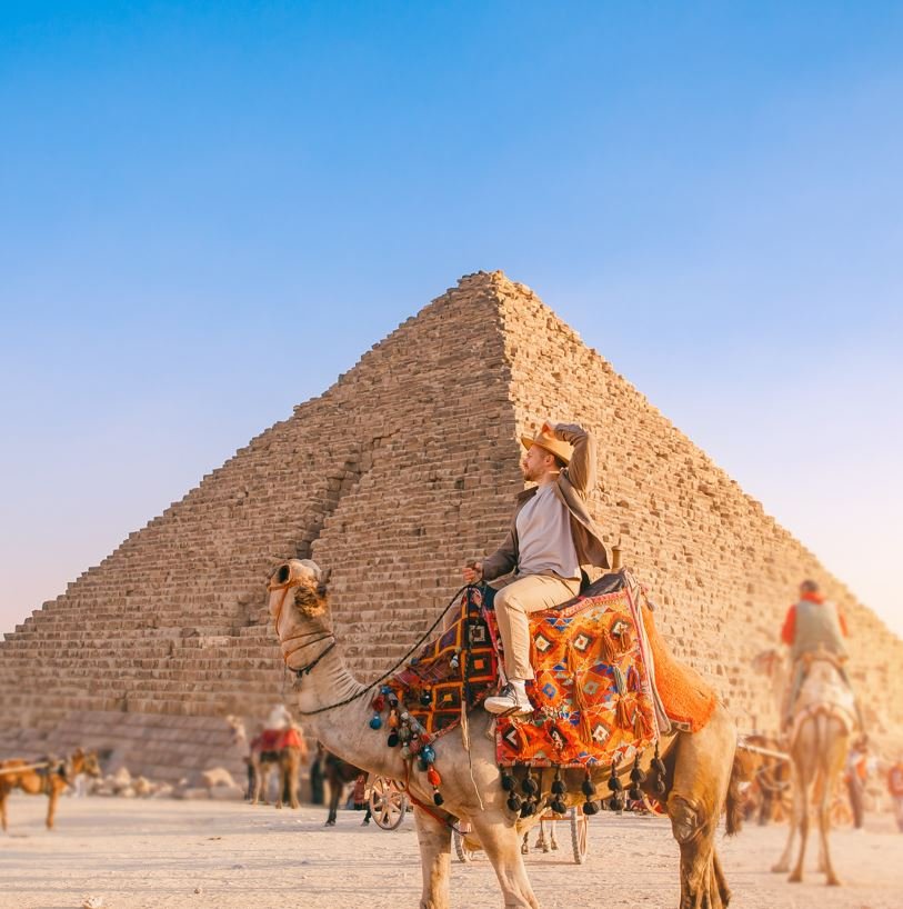 Happy-Tourist-man-with-hat-riding-on-camel-background-pyramid-of-Egyptian-Giza-sun-light-Cairo-Egypt