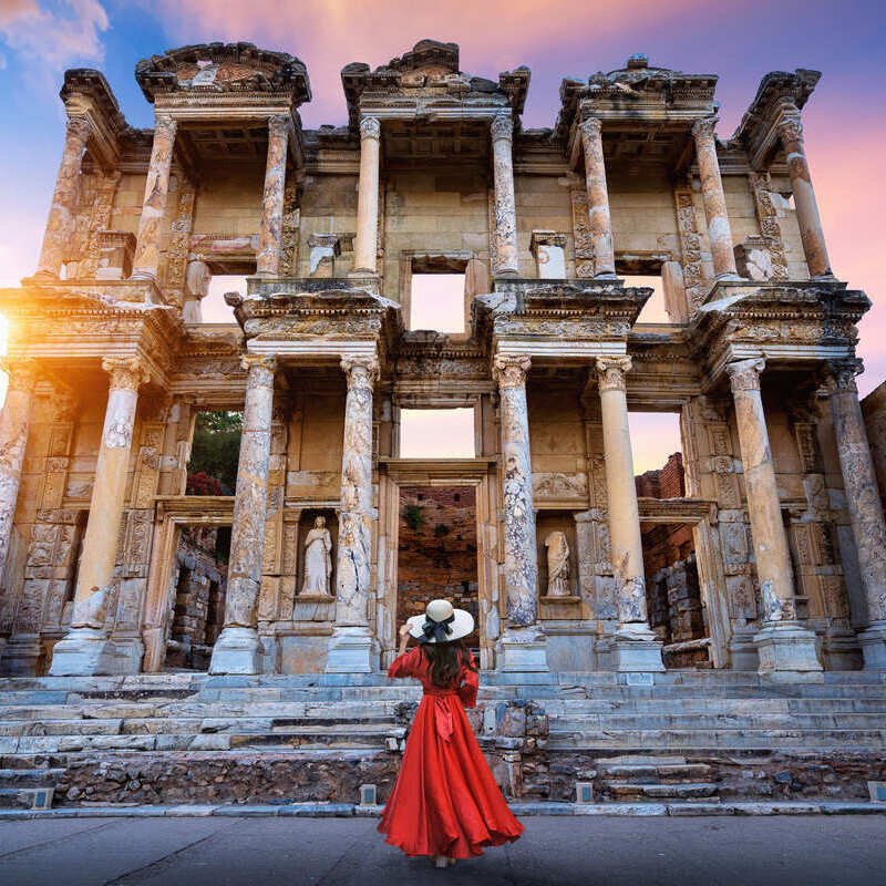 Young Female Tourist Posing By The Facade Of The Library Of Celsus In The Ancient City Of Ephesus, Near Selcuk, Turkey, U.S. dollar goes furthest here