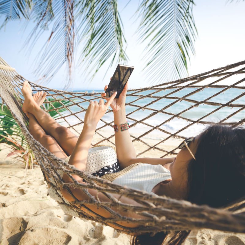 Summer vacations concept, Happy woman with white bikini, hat and sunglass using mobile phone in hammock on tropical beach at sunset, Koh mak, Thailand