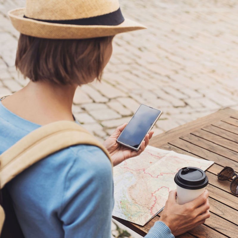 Young traveller woman sitting in a cafe terrace and planning trip with map and smartphone. Travel and active lifestyle concept