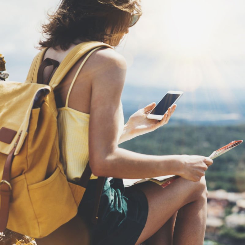 Young girl with bright backpack looking at phone and map. Said view tourist traveler on background mountain, blue sea, sun flare. 