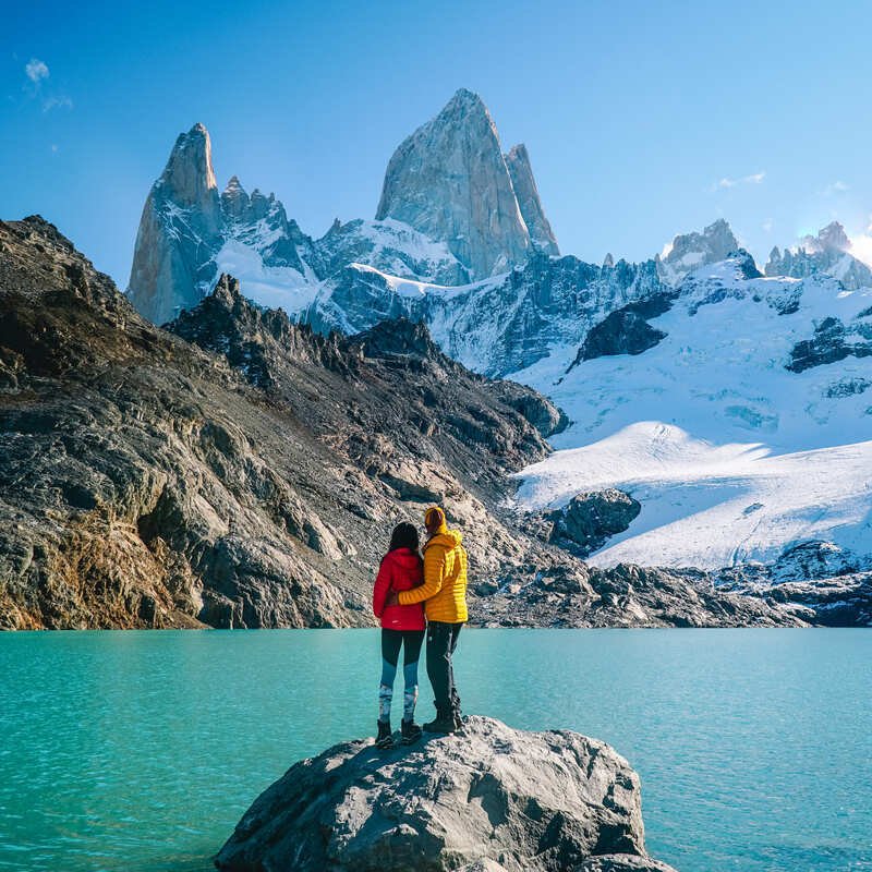 Couple Embracing As They Stan On A Rock By The Shores Of A Glacial Lake, Surrounded By Alpine, Snow Capped Peaks In Patagonia, Southern Argentina, South America