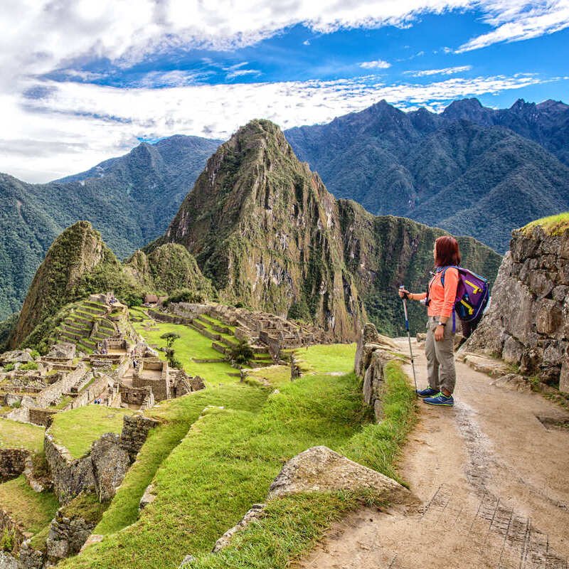 A Female Tourist Leaning On A Trekking Pole As She Admires The View Of Machu Picchu Surrounded By Green Peaks In Peru