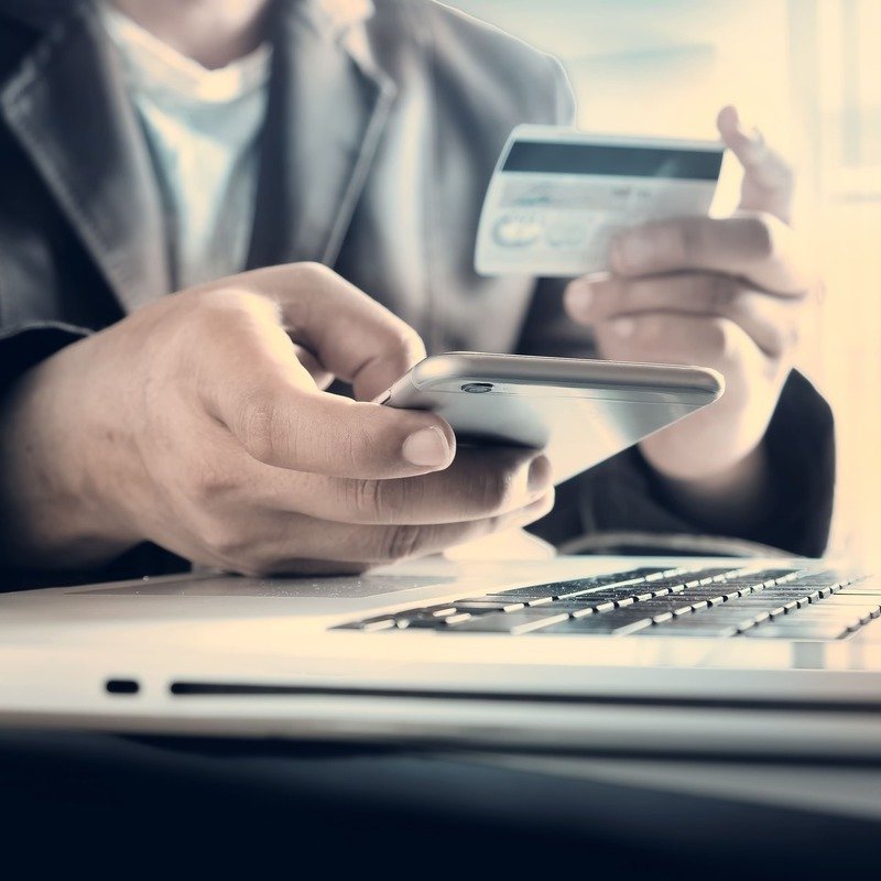 A Male Person Making An Online Payment As He Checks His Credit Card Information And Looks At His Phone And Laptop