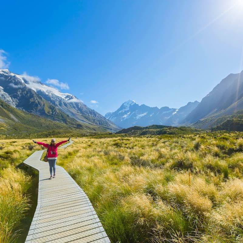 Female Solo Traveler Standing Over Footpath Leading To Snow Capped Mountains In New Zealand