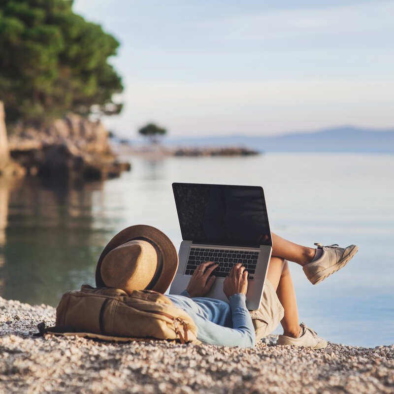 Digital Nomad Lying On His Rucksack As He Works From His Laptop On A Pebbly Beach In An Unspecified Location