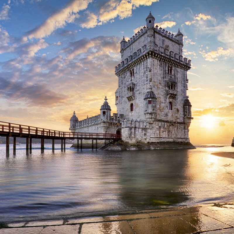 The Belem Tower over the Tagus River in Portugal. A huge tower sits on the beach with a bridge stretching out. 