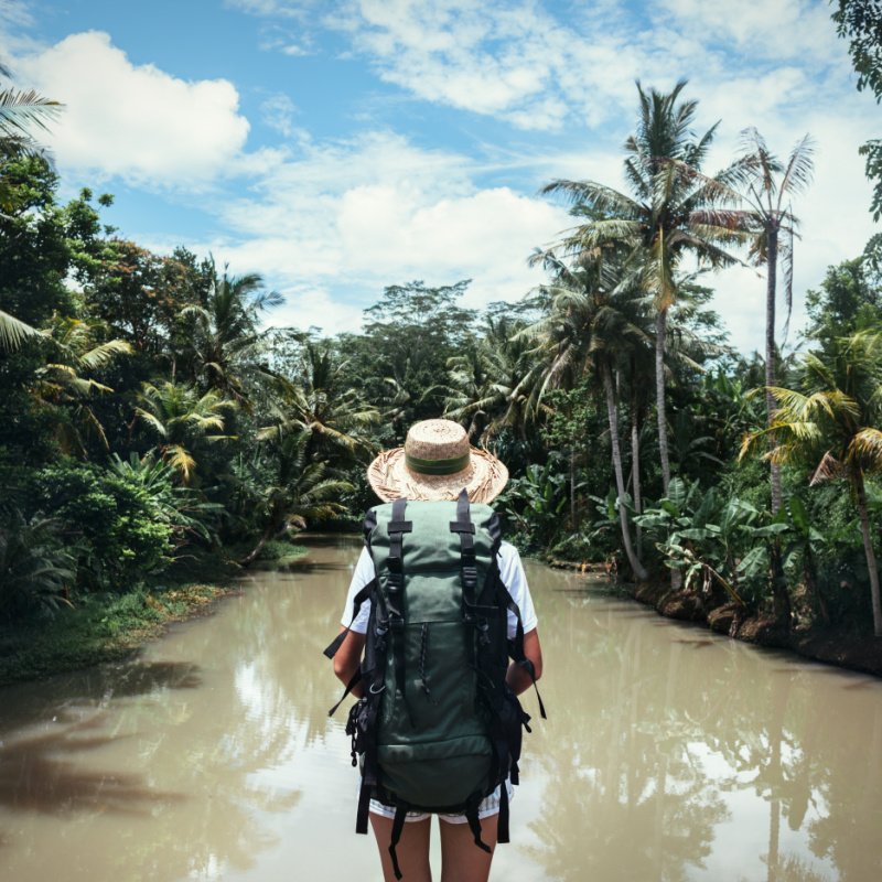 Traveling woman with backpack and straw hat looking at tropical river at sunny day.