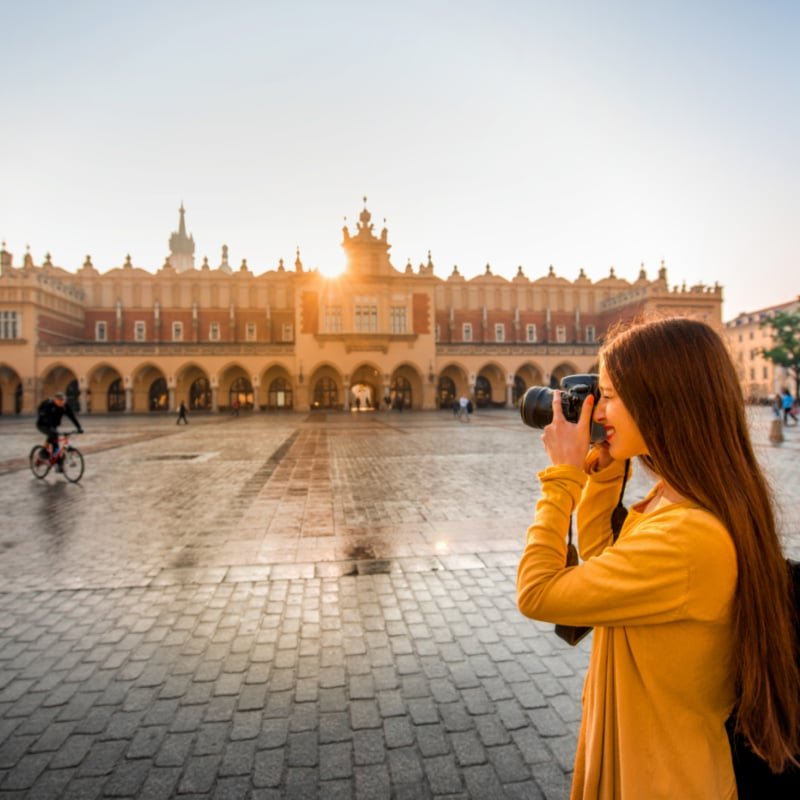 Young female tourist with camera and backpack photographing Cloth Hall in the old city center of Krakow (1)