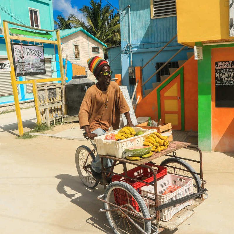 A Beach Vendor Pushing A Wheeled Street Food Stall Down The Colorful Streets Of Caye Caulker, A City In Belize, Central America