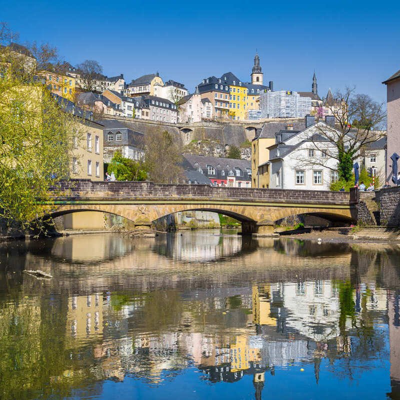 View Of An Old Stone Bridge In The Grund Distruct Of Luxembourg City, With Luxembourg Castle And The Chemin Of La Corniche For Backdrop, Western Europe