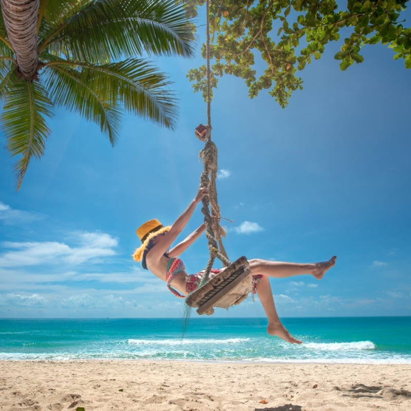 Solo Female Traveler Swings From A Palm Tree On An Island In Thailand, Southeast Asia