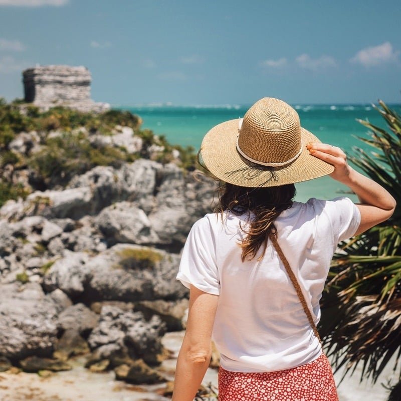 A Female Tourist Holding Her Straw Hat On Her Head As She Admires The Tulum Ruins, Mexican Caribbean, Mexico