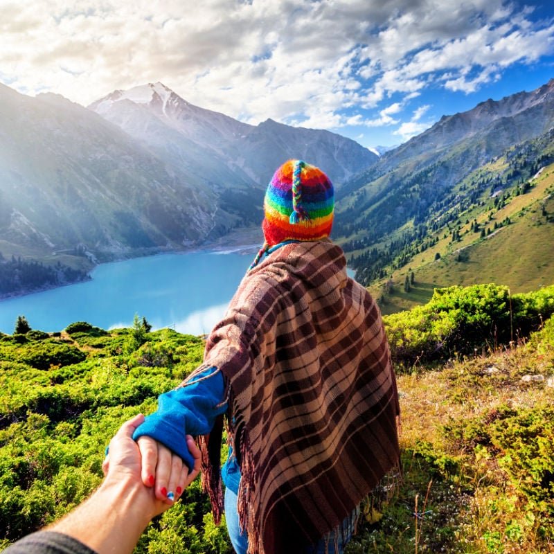 Tourist woman in rainbow hat and brown poncho holding man by hand and going to the lake in the mountains in peru