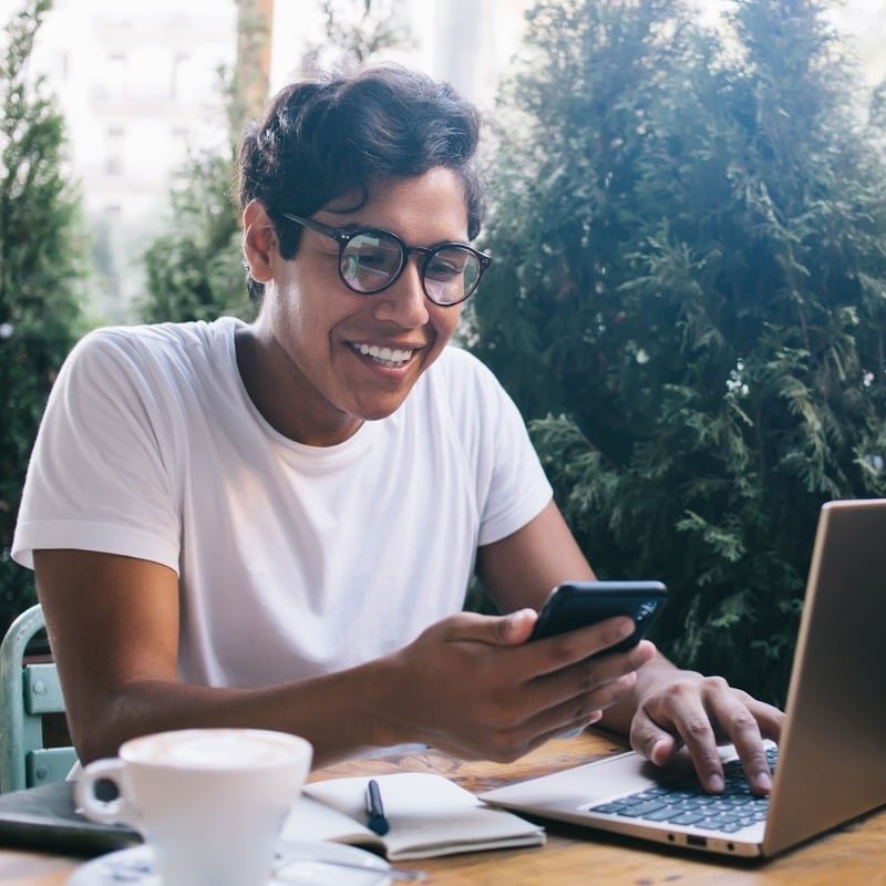 Smiling Remote Worker, Digital Nomad Checking His Phone As He Works From His Computer In A Cafe Setting, Unspecified Location