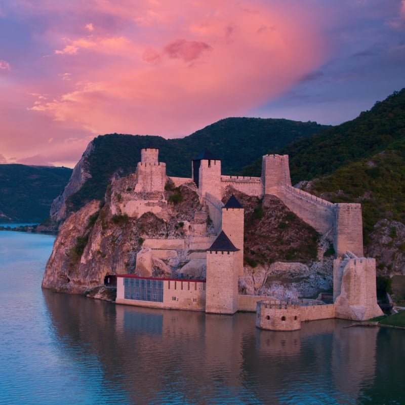 Aerial, across lake view of the medieval fortress Golubac over  Danube river. Fortress towers illuminated by pink light. Sunset, pink and red clouds sky. Outdoor and traveling theme. Serbia.