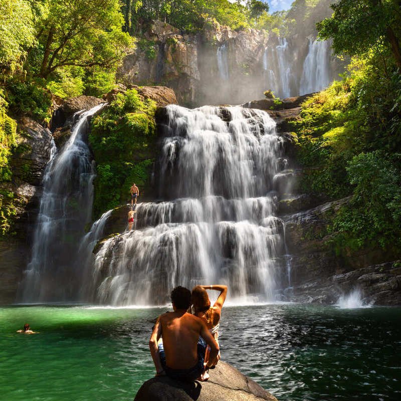 Couple Of Travelers Sat On A Rock As They Admire A Waterfall In Manuel Antonio National Park In Costa Rica, Central America