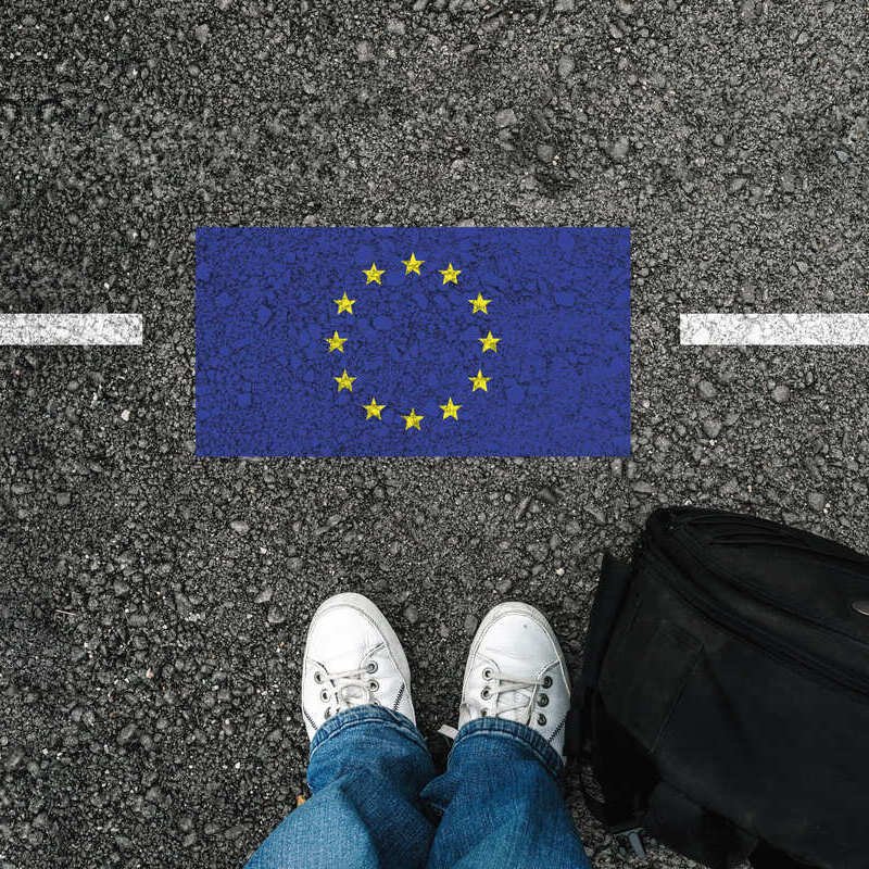 A Person Stands Behind A White Line And An European Union EU Flag Painted On The Ground Alongside Their Backpack